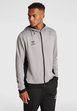 Joven con sudadera gris con cremallera con hombros con puntos y pantalones deportivos negros, de pie con una mano en el bolsillo mirando hacia la izquierda.