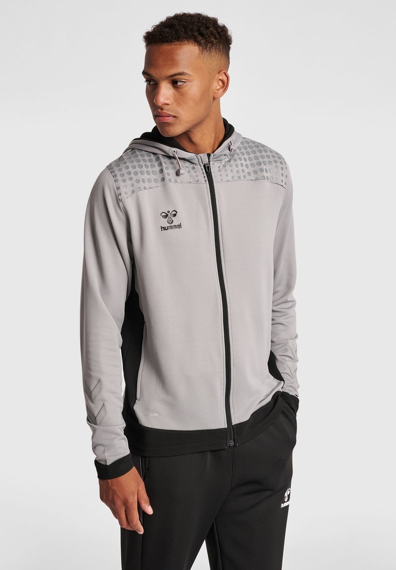 Młody mężczyzna w szarej bluzie z kapturem na zamek z kropkami na ramionach i czarnych spodniach dresowych, stojący z jedną ręką w kieszeni, patrzący w lewo.