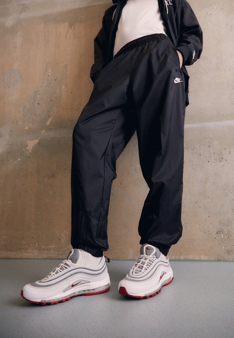 Osoba oblečená v černých Nike teplákových kalhotách a bílých Nike Air Max teniskách s červenými akcenty, stojící proti texturované betonové zdi.