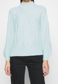 Ljust blå stickad tröja med hög hals, strukturerade mönster och ribbade muddar, i avslappnad passform och med intrikata kabeldetaljer.