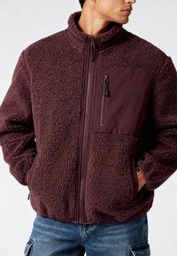 Giacca in pile bordeaux con cerniera frontale, collo alto e tasca sul petto con zip. Tessuto strutturato con finitura soffice e morbida.