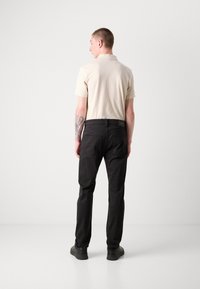Beige poloshirt en zwarte broek, met een slim fit ontwerp, zichtbare tatoeage op de arm en zwarte schoenen. Eenvoudige, duidelijke lijnen.