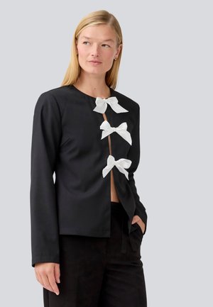 Top nero a maniche lunghe con tre fiocchi bianchi decorativi all'apertura frontale. Tessuto morbido e design a taglio dritto. Abbinato a pantaloni neri.