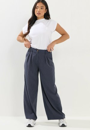 Kobieta w białym topie bez rękawów i granatowych szerokich spodniach w drobne prążki stojąca, regulująca pasek rękoma.