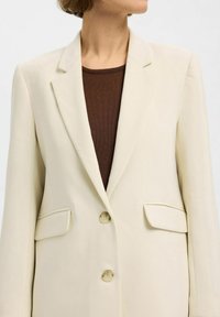 Beige skræddersyet blazer med spidse lapeller, to forlommer og to knapper. Båret over en brun top, som viser en glat tekstur.