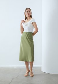 Haut blanc à épaules nues avec des plis et un détail floral, associé à une jupe midi en satin vert olive et des talons beiges à lanières.