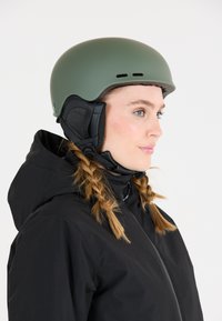 Matte olijfgroene skihelmet met ventilatiesleuven, zwarte oorbedekkingen en een verstelbare kinband. Gepaarde met een zwarte jas.