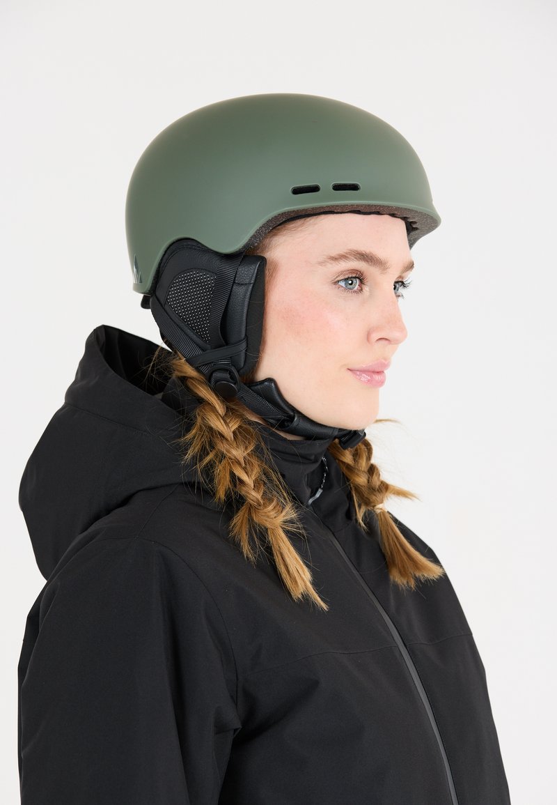 Matte olijfgroene skihelmet met ventilatiesleuven, zwarte oorbedekkingen en een verstelbare kinband. Gepaarde met een zwarte jas.