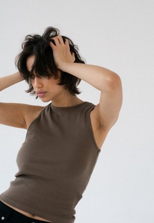 Kobieta o krótkich ciemnych włosach, ubrana w bezrękawnik w kolorze brązowym, trzymająca głowę obiema rękami na jednolitym jasnym tle.