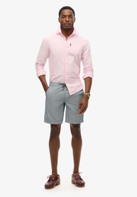 Chemise à boutons rose clair, shorts rayés gris, chaussures en cuir marron avec lacets. Le modèle se tient avec les mains dans les poches, sur un fond neutre.