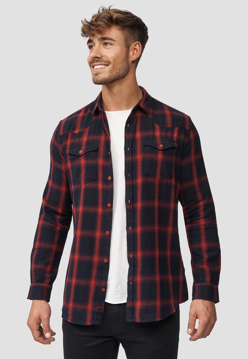 Camisa de franela de cuadros rojos y marinos con mangas largas, dos bolsillos en el pecho y cierres de botones. Textura suave con un corte relajado.