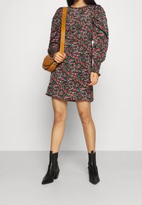 Kleid mit floralem Muster und langen Puffärmeln, schwarzer Hintergrund und leicht ausgestelltem Saum. Kombiniert mit schwarzen Stiefeletten und einer braunen Tasche.