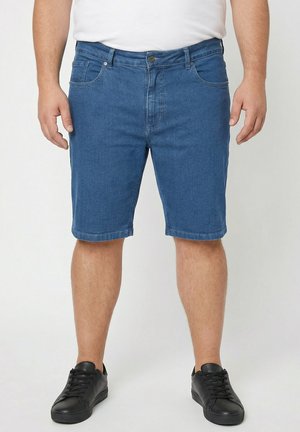 Mand iført blå cowboyshorts, sorte sneakers og en hvid skjorte, stående mod en ensfarvet lys baggrund.