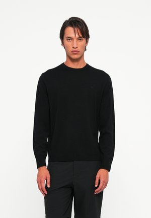Jonge man die recht vooruit staat, gekleed in een zwarte lange mouwen trui en zwarte broek, tegen een effen witte achtergrond.