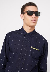 Námořnická košile na knoflíky s malými bílými a žlutými čtvercovými vzory, obsahující náprsní kapsu se žlutým akcentem. Celý look doplňují černé sluneční brýle.