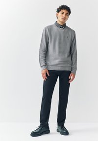 Grå ribbstickad tröja med rund halsringning, lager-på-lager över en svart skjorta med prickar, kombinerad med smala svarta byxor och mörka skor.