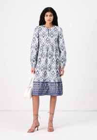 Rochie cu mânecă lungă, albă și albastră, cu un model floral ornate. Are un decolteu rotund și o fustă în clape. Modelul ține o geantă mică.