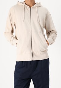 Felpa con zip in cotone beige, dotata di cappuccio con cordino, tasche frontali e polsini a coste. Indossata con pantaloni blu scuro. Texture morbida.