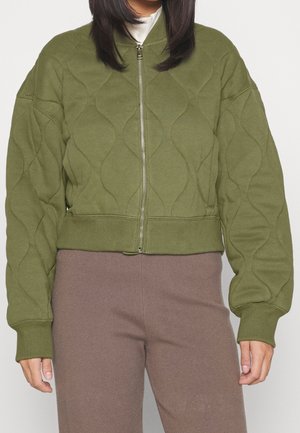 Femme portant une veste matelassée zippée vert olive avec poignets et ourlet côtelés sur un pantalon mauve, debout sur un fond uni.