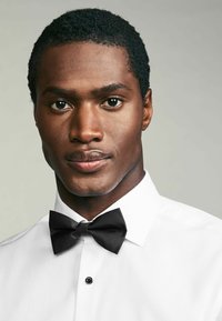 Chemise blanche avec un tissu texturé et un nœud papillon à rayures noires, comportant un seul bouton noir au col. Gros plan sur un modèle masculin.
