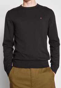 Maglione nero lavorato a maglia con scollo rotondo, polsini e orlo a costine, decorato con un piccolo logo tricolore sul petto. Abbinato a pantaloni color khaki.