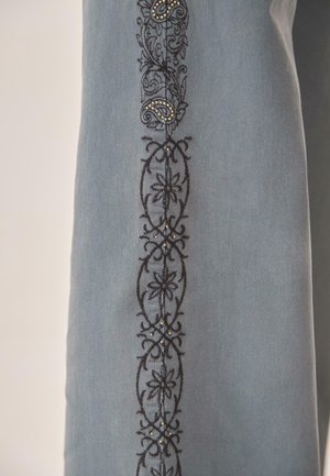 Tela azul claro con bordado floral y de paisley negro adornado con pequeñas tachuelas metálicas en un patrón vertical.