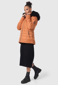 Orange insulated jacket with a faux fur hood, quilted design, zippered pockets, paired with a black ribbed midi skirt and chunky black boots.