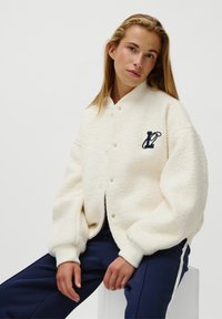 Kermanvärinen fleece-bomberi takki, jossa on ribbaattu kaulus ja hihansuut, ja vasemmassa rinnassa on laivastonsininen brodeerattu logo.