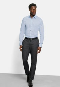 Hellblaues, gemustertes Hemd mit langen Ärmeln, kombiniert mit dunkelgrauen Hosen und schwarzen Schuhen, vor einem weißen Hintergrund stehend.