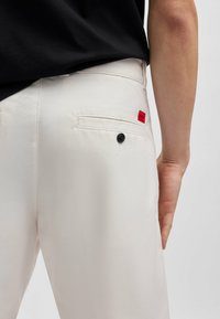 Pantalons beige clair avec une texture lisse, dotés d'une poche arrière avec un bouton noir et une petite étiquette de marque rouge.