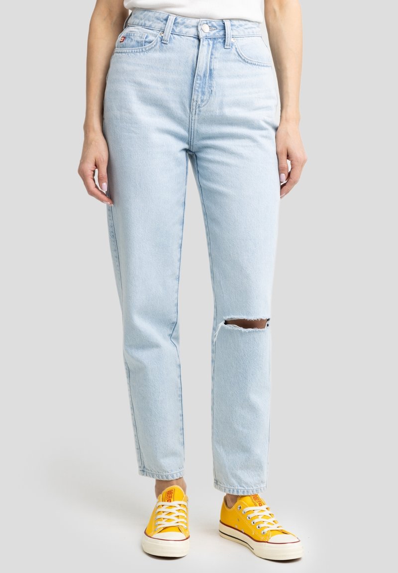 Jean taille haute bleu clair avec une déchirure au niveau du genou, porté avec des baskets basses jaune vif et un haut blanc sur un fond gris.