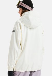 Witte waterdichte jas met een capuchon, elastische manchetten en een zwart logo-opdruk op de mouw. Gecombineerd met een zwarte beanie en skibril.