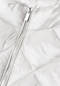 Gros plan sur une veste matelassée blanche montrant une fermeture éclair argentée centrée et des détails de couture rembourrée.