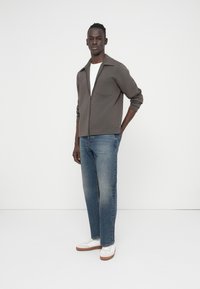 Γκρι ζακέτα με φερμουάρ και γιακά με μυτερή άκρη, σε συνδυασμό με ανοιχτό μπλε τζιν με ίσια εφαρμογή και λευκά αθλητικά παπούτσια με καφέ σόλες.