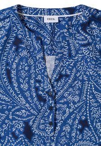 Chemise bleue à boutons avec motif abstrait paisley blanc et boutons noirs, avec un col rond et une étiquette "CECIL" à l'intérieur du col.