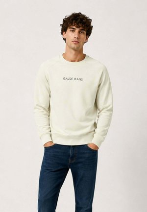 Giovane uomo con capelli ricci che indossa una felpa crema con la scritta "GAUDÍ JEANS" e jeans blu scuro, mani nelle tasche, sfondo neutro.