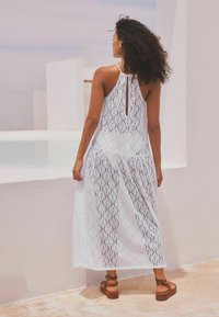 Robe maxi blanche avec un motif en dentelle, dos nu et décolleté halter. Associée à des sandales à plateforme marron. Texture lisse.