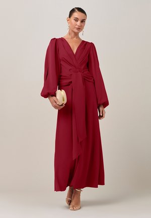 Femme portant une robe bordeaux longue jusqu'au sol avec de longues manches bouffantes et un profond décolleté en V, tenant une pochette dorée et portant des talons beiges.