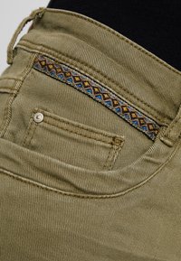 Khaki farkut, joissa on teksturoitu kangas, ja koristeellinen broderad tasku sinisin ja ruskein geometrisin kuvioin.