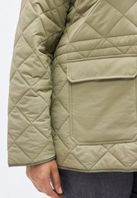 Πράσινο μπουφάν με καπιτονέ φινίρισμα και υφή, διαθέτει μία τσέπη με μπαλώματα και ίσια ρ ίγα στο κάτω μέρος, τονίζοντας γεωμετρικά μοτίβα ρομβοειδούς σχήματος.