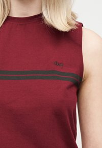 Μπλούζα χωρίς μανίκια σε χρώμα μαρόν με δύο σκούρες οριζόντιες ρίγες και μικρό λογότυπο "obey" στο πάνω μέρος του στήθους, φορεμένη από ένα άτομο με ανοιχτό ξανθό μαλλί.