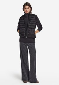Schwarze Steppweste mit horizontalem Rauten-Muster, mit Reißverschluss, über einem schwarzen Rollkragenpullover getragen. Kombiniert mit weit geschnittenen, dunkelgrauen Jeans und Stiefeletten.