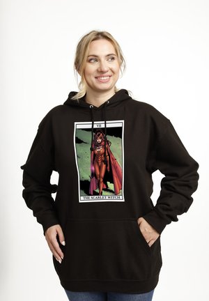 Uśmiechnięta kobieta w czarnej bluzie z kapturem z grafiką karty tarota Scarlet Witch, ręce w kieszeniach, stojąca na jednolitym białym tle.