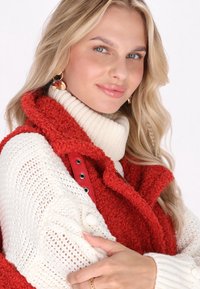 Rote, flauschige Weste mit hohem Kragen, über einem dicken, cremefarbenen, gestrickten Pullover mit strukturierten Akzenten getragen. Goldene Ohrringe verleihen das Detail.