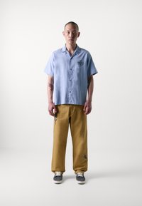 Camicia azzurra a maniche corte con logo frontale, abbinata a pantaloni beige con design ricamati. Sneakers grigie completano il look.