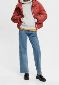 Giacca puffer trapuntata rossa sopra un felpa grigia con cappuccio, abbinata a jeans a gamba larga blu e stivali neri. Presenta un cappuccio e una chiusura a zip.