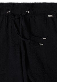 Pantalon noir texturé avec une taille élastique, cordon noir avec embouts argentés et une petite étiquette de marque en métal près de la poche.