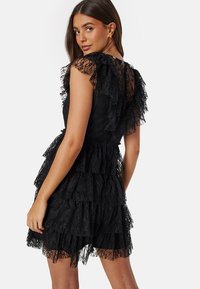 Robe en dentelle noire avec des volants superposés, une taille cintrée et un détail en trou de serrure au dos avec des fermetures à bouton. La dentelle texturée ajoute de la profondeur.