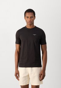 Zwarte katoenen T-shirt met korte mouwen en een ronde hals. Heeft een klein wit logo op de borst. Draagt met lichtgekleurde shorts.