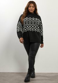 Suéter de punto negro con patrones geométricos y florales en blanco, cuello alto y dobladillo acanalado, combinado con leggings de cuero sintético negro y botas.